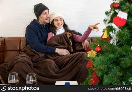 Smiling couple lover in sweater enjoying and talking on sofa. Woman pointing to outside while celebrating with glasses of red wine in living room. Romantic winter holiday and Christmas eve together concept