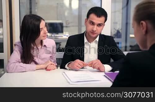 Smiling couple signing house purchase agreement with real estate agent in light modern office. Cheerful young man in formalwear studying the contract and signing the document after consulting with his wife.