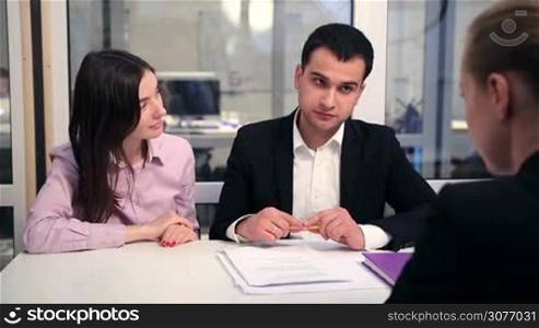Smiling couple signing house purchase agreement with real estate agent in light modern office. Cheerful young man in formalwear studying the contract and signing the document after consulting with his wife.