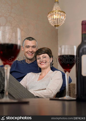 Smiling couple sitting at table in living room wine glasses on foreground (portrait)