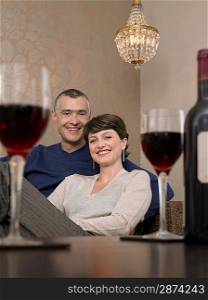 Smiling couple sitting at table in living room wine glasses on foreground (portrait)
