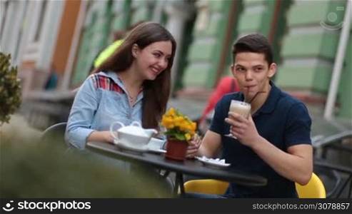 Smiling couple sitting at the table in outdoors cafe and talking. Romantic couple enjoying leisure, drinking coffee and tea in sidewalk cafe on summer day. Joyful hipster couple relaxing at coffee shop during romantic date.