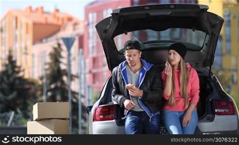 Smiling couple sitting in trunk of modern car and having a snack during moving in to their new apartment.