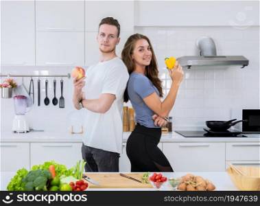 Smiling couple standing in kitchen while preparing healthy food. Man holding red apple and woman holding yellow bell pepper.