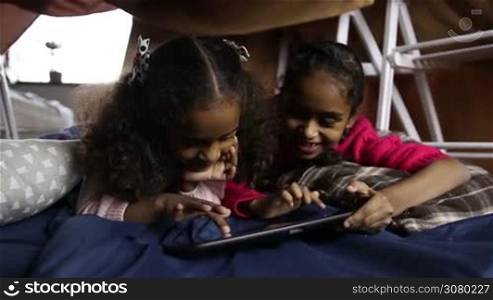 Smiling cute african american sisters scrolling pages on digital tablet while lying on the floor in cubby house made of blankets and chairs. Positive mixed race little girls playing online game using touchpad at home. Dolly shot. Slow motion.
