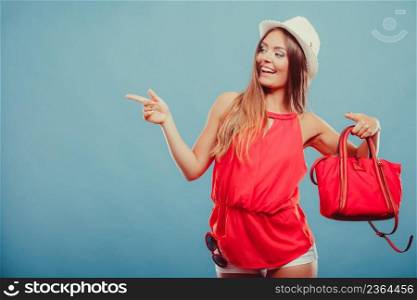 Smiling cute attractive young woman girl in red shirt and hat with handbag and sunglasses in studio on blue showing blank empty copyspace. Summer female fashion  and advertisement.. Fashion woman showing blank empty copyspace.