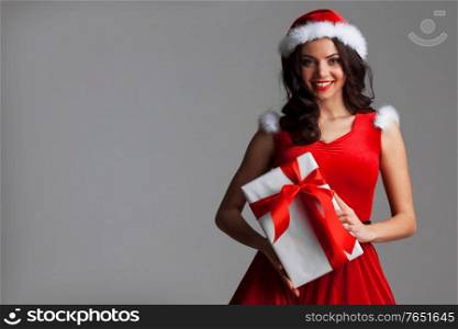 Smiling cute girl in red christmas outfit holding gift box on gray background. Girl with christmas gift box