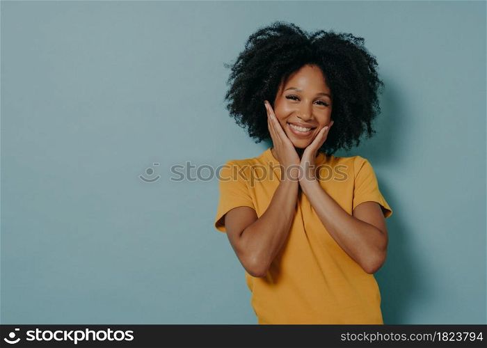 Smiling cute mixed race young woman in yellow tshirt touching cheeks with hands and feeling happy after hearing compliment from boyfriend, standing isolated on blue background with copy space. Cute mixed race young woman touching cheeks with hands and feeling happy after hearing compliment