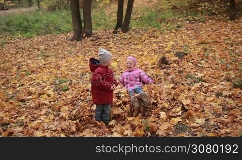 Smiling cute toddler boy in warm clothes and his attractive teenage sister in eyeglasses jumping in large pile of autumn leaves in the park. Joyful kids playing with yellow fallen leaves, throwing them up into the air on a warm fall day. Slow motion.