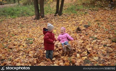 Smiling cute toddler boy in warm clothes and his attractive teenage sister in eyeglasses jumping in large pile of autumn leaves in the park. Joyful kids playing with yellow fallen leaves, throwing them up into the air on a warm fall day. Slow motion.