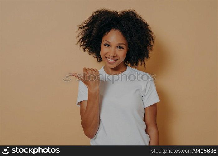 Smiling dark-skinned woman with curly hair, pointing aside with thumb, indicating blank space for promotion. Isolated on beige wall.