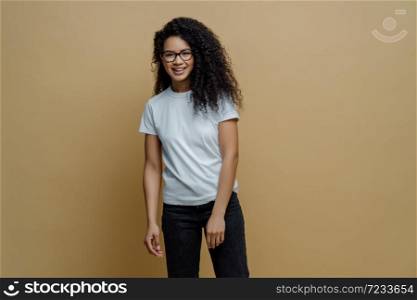 Smiling dark skinned young woman with glad face expression, wears optical glasses, white t shirt and jeans, poses carefree against beige background. People, happy mood and lifestyle concept.