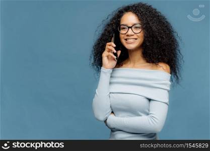 Smiling delighted Afro American woman with curly hair, talks on smart phone, discusses some pleasant moments after party, looks happily into distance, wears glasses and jumper, isolated on blue wall