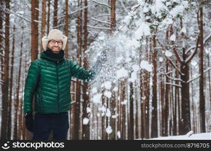 Smiling delightful male dressed in warm clothes, stands in winter forest, throws snow in air, has fun alone, has good mood, expresses positive emotions and feelings. Positiveness concept