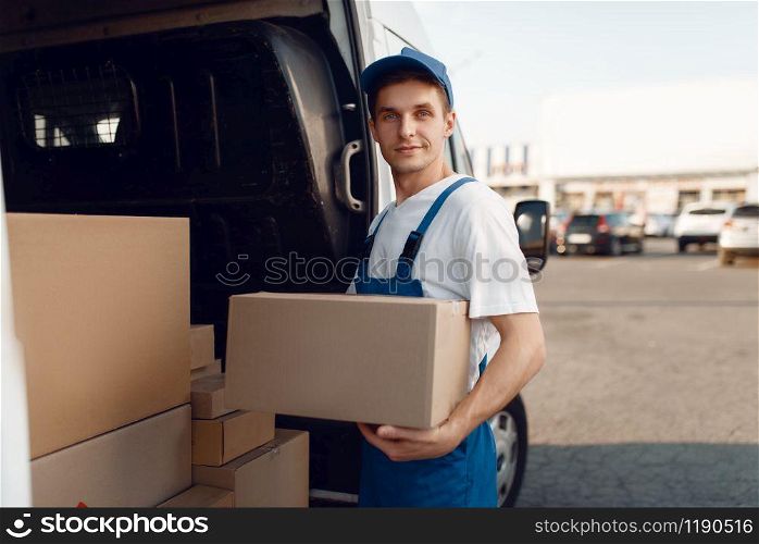 Smiling deliveryman with carton box at the car, delivery service. Man in uniform holding cardboard package, male deliver, courier job. Smiling deliveryman with carton box at the car