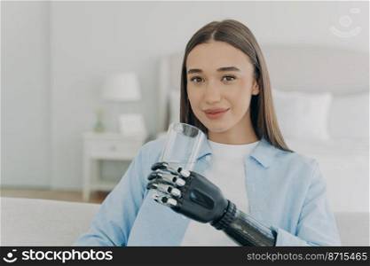 Smiling disabled girl holding glass with clean water by bionic prosthetic arm. Happy young woman holds drink with artificial robotic hand looking at camera. Disability and daily routine concept.. Smiling disabled girl holds glass with water by bionic prosthetic arm. Disability, healthy lifestyle