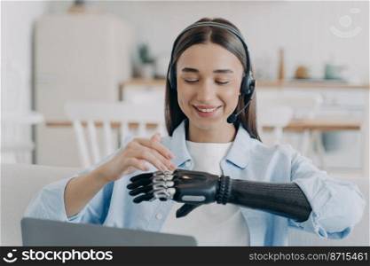 Smiling disabled girl student with bionic prosthesis instead of arm wearing headset sitting at laptop, setting robotic hand, learning or working online. Lifestyle of people with disabilities.. Smiling disabled girl student in headset using bionic prosthetic arm sitting at laptop at home