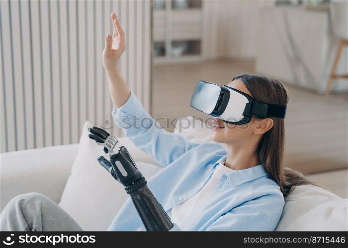 Smiling disabled girl wearing VR glasses interacting with virtual reality objects by bionic prosthetic arm, sitting on sofa. Young woman training to use artificial limb at home. Medical high tech.. Smiling disabled girl in VR glasses interacts with virtual reality objects by bionic prosthetic arm