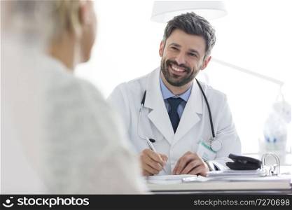Smiling doctor discussing with mature patient while writing prescription at desk in hospital