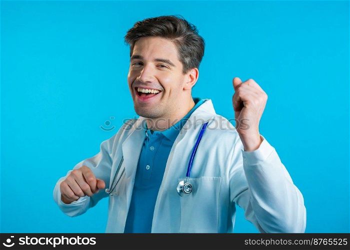 Smiling doctor funny dancing, success and luck at work. Young handsome doc man in professional medical white coat is isolated on blue studio background. High quality photo. Smiling doctor funny dancing, success and luck at work. Young handsome doc man in professional medical white coat is isolated on blue studio background.