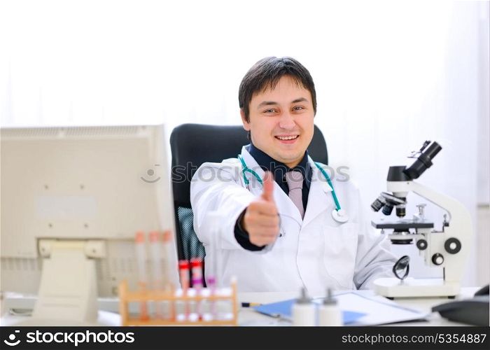 Smiling doctor sitting at office table and showing thumbs up gesture&#xA;