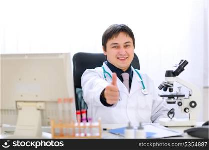 Smiling doctor sitting at office table and showing thumbs up gesture&#xA;
