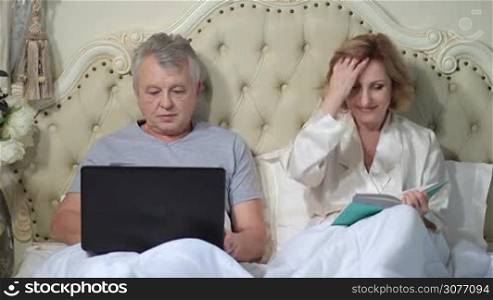 Smiling elderly husband and wife relaxing in bed with laptop and book. Relaxed senior couple lounging in family bed in the morning. Handsome husband using laptop while his attractive wife reading an interesting book in the bedroom at home.
