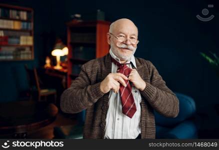 Smiling elderly man puts on a tie in home office. Bearded mature senior in living room, old age businessman. Elderly man puts on a tie in home office