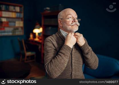 Smiling elderly man puts on a tie in home office. Bearded mature senior in living room, old age businessman. Elderly man puts on a tie in home office
