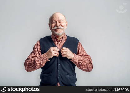 Smiling elderly man with mustache puts on a jacket, grey background. Cheerful mature senior looking at camera in studio. Smiling elderly man with mustache puts on a jacket