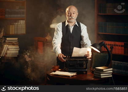 Smiling elderly writer works on retro typewriter in home office. Old man in glasses writes literature novel in room with smoke, inspiration. Smiling elderly writer works on retro typewriter