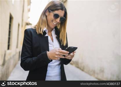 smiling elegant young woman using smartphone buildings street