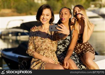 Smiling ethnic woman taking self portrait on cellphone with black girlfriend showing tongue and blonde making grimace on pier. Funny diverse women taking selfie on smartphone on seafront