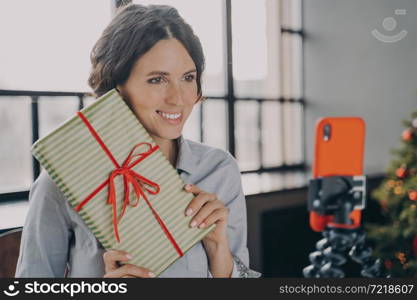 Smiling European female blogger streaming live online via mobile phone on tripod holding decorated gift and telling how to wrap Xmas presents better and easier. New Year and Christmas holiday concept. Smiling female blogger streaming live online via mobile phone on tripod during Christmas holidays