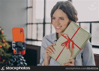 Smiling European female blogger streaming live online via mobile phone on tripod holding decorated gift and telling how to wrap Xmas presents better and easier. New Year and Christmas holiday concept. Smiling female blogger streaming live online via mobile phone on tripod during Christmas holidays