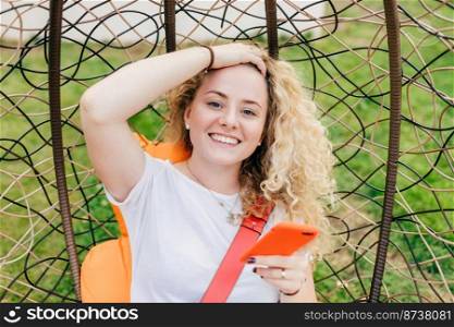 Smiling European female with broad smile, curly hair, dressed in casual white t shirt, uses modern electronic device for surfing social networks, happy to recieve message on invitation to party