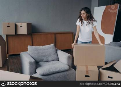 Smiling european woman is packing things. Girl moves to new house. Young lady is unpacking cardboard boxes in new apartment. Big new living room with armchair. Real estate purchase concept.. Smiling european woman is packing things. Young lady is unpacking cardboard boxes in new apartment.