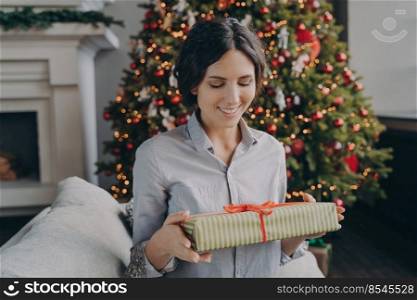 Smiling european woman sitting on sofa in room decorated for xmas holidays holding wrapped Christmas present, happy hispanic female looking at gift box with red bow while celebrating New Year at home. Smiling european woman sitting on sofa in room decorated for xmas holidays holding Christmas present