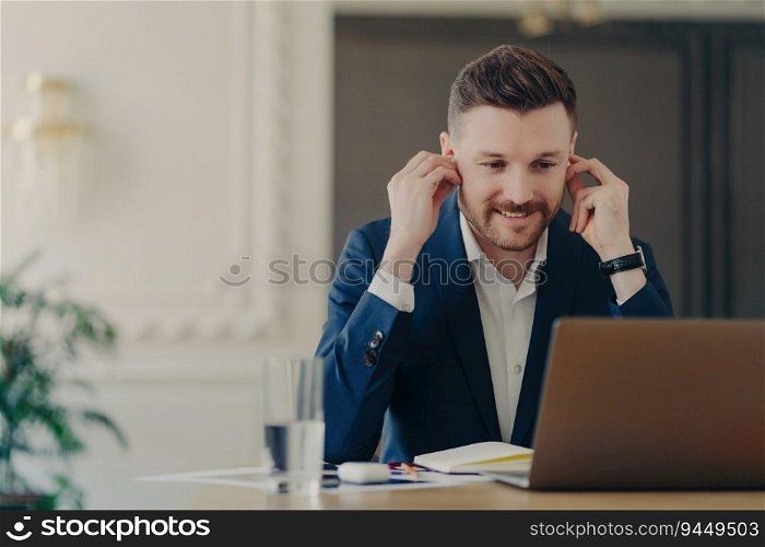 Smiling executive manager in blue suit, online meeting with employees, wireless earphones, sitting at office desk with laptop.
