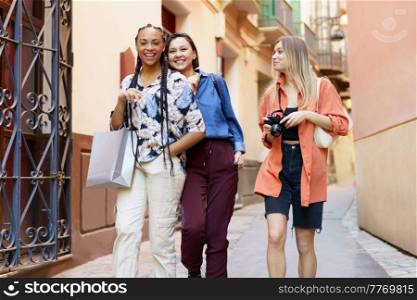 Smiling fair haired woman with camera looking at cheerful diverse female friends while going home after buying new clothes. Joyful multiracial females strolling together after shopping