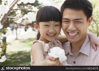 Smiling father and daughter enjoying the cherry blossoms on the tree in the park in springtime, daughter holding a flower