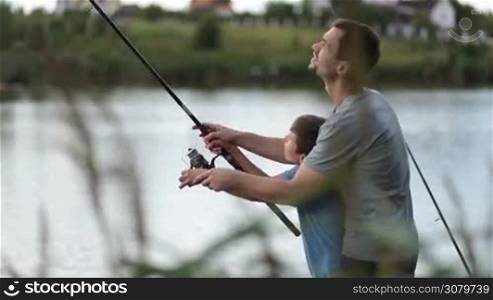 Smiling father and joyful teenage boy spending great time together fishing with rod on the lake. Side view. Foreground blurry canes flying in the wind. Positive dad and son angling with fishing rod at freshwater pond while enjoying leisure in summer.
