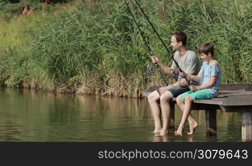 Smiling father and teenage boy sitting on wooden pier swung with their legs in water while fishing together by the lake in summer day. Positive dad communicating with son while fishing with rods near pond over beautiful landscape background. Slow motion.