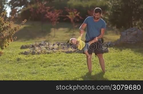 Smiling father swinging his giggling infant son by hands upside down in public park against beautiful summer landscape background. Handsome dad playing and having fun with cute toddler baby boy as they enjoy time and leisure together in nature.
