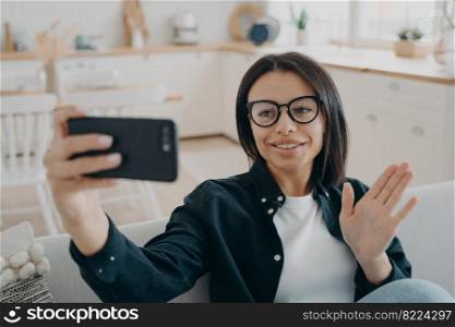 Smiling female answers video call holding smartphone, greeting waving hand hello, sitting on sofa at home. Modern young woman blogger wearing glasses chatting online, welcoming followers.. Female in glasses answers video call holding smartphone, greeting waving hand hello, sitting at home
