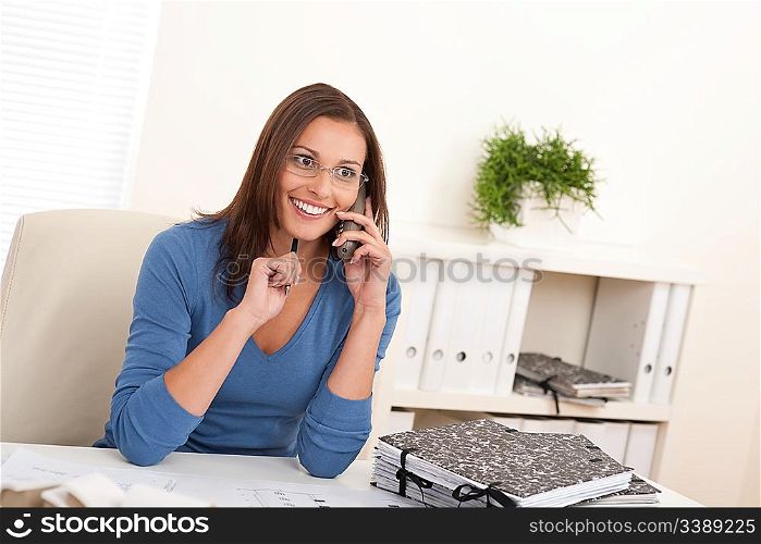 Smiling female architect holding phone and pen sitting at the office