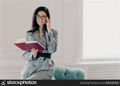 Smiling female business reporter in formal clothes holds opened notebook and pen, writes down information or checklist, wears formal clothes, works remotely from home, prepares for formal meeting