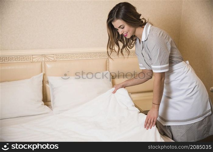 smiling female chambermaid making bed hotel room