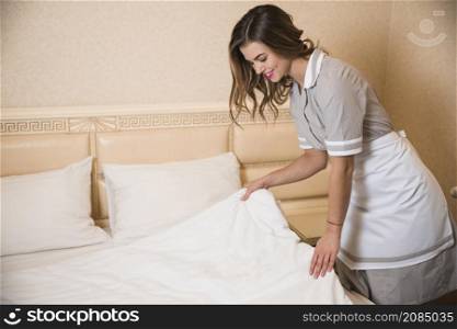smiling female chambermaid making bed hotel room