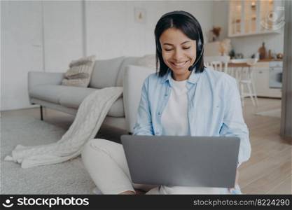 Smiling female coach wearing headset working on laptop conducts online lesson, sitting on the floor at home. Friendly woman in wireless headphones listening webinar. Distance education, e-learning.. Smiling female coach in headset work on laptop or study online, sitting on floor at home. E-learning
